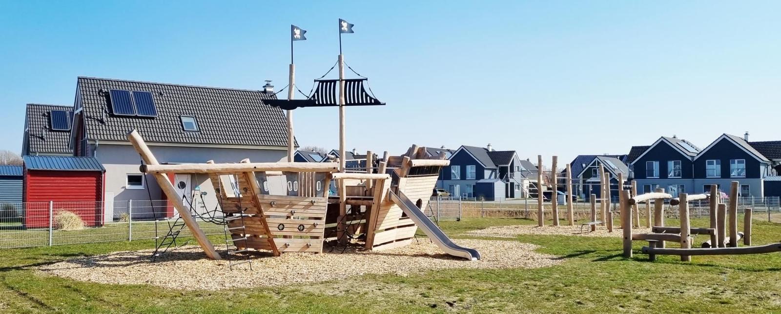 Holzschiff mit Rutsche und Masten im Garten. Hintergrund: Wohnhäuser und klarer Himmel.