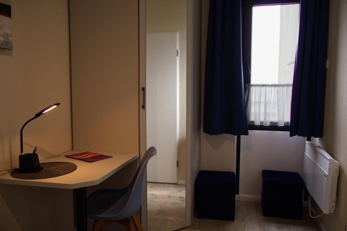 Schreibtisch mit Lampe, Stuhl, Fenster mit Vorhängen und zwei Hocker.