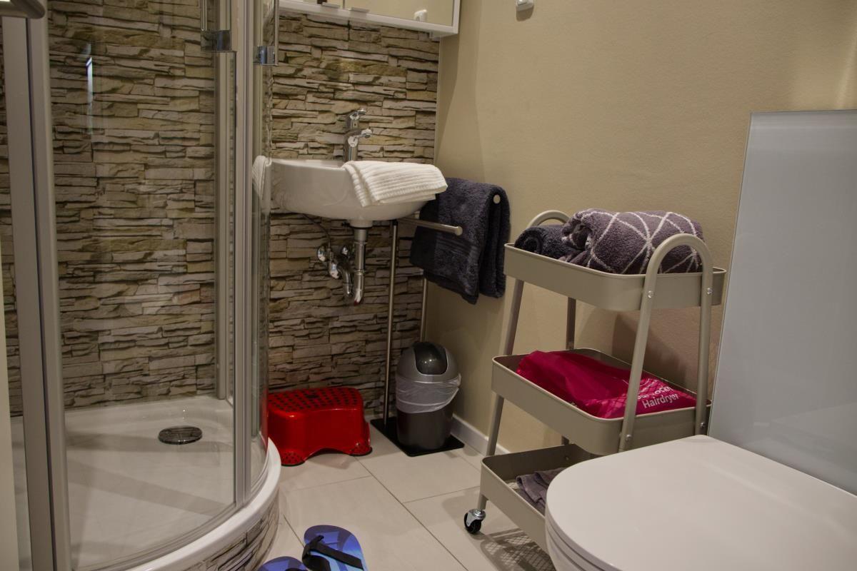 Badezimmer mit Dusche, Waschbecken und Toilette. Auf Wagen liegen Handtücher und ein Haartrockner.