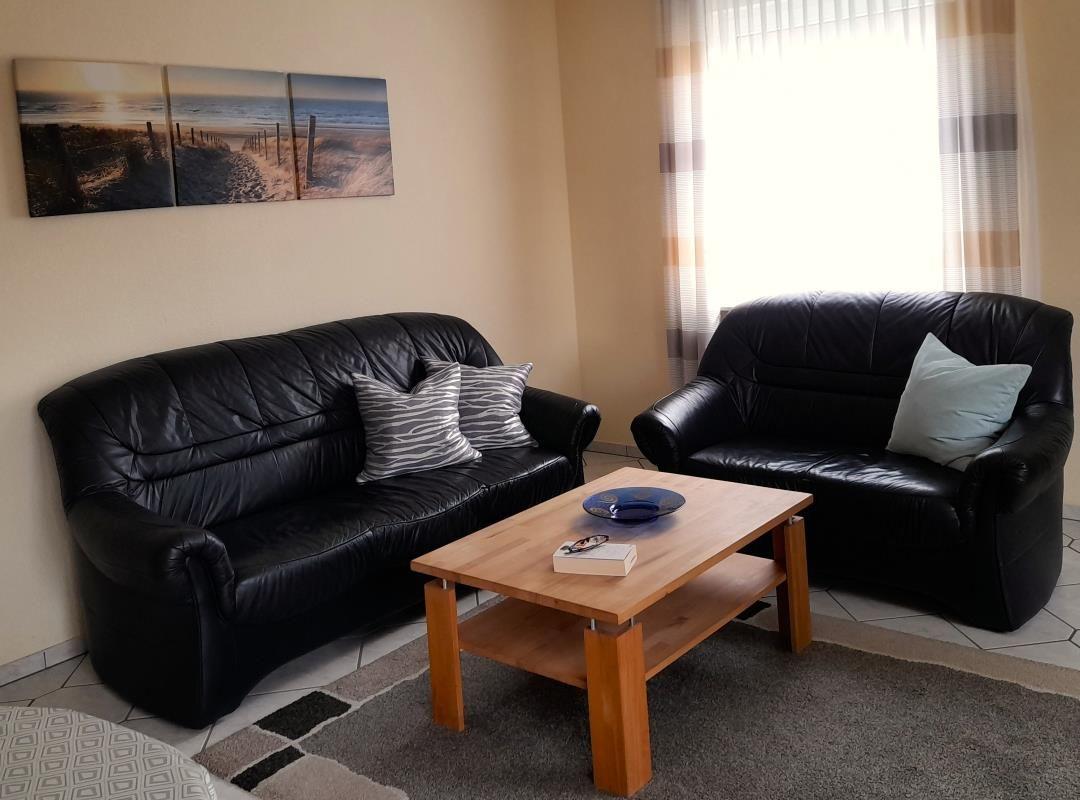 Two black sofas, wooden table, wall art with beach motif, window with curtains.