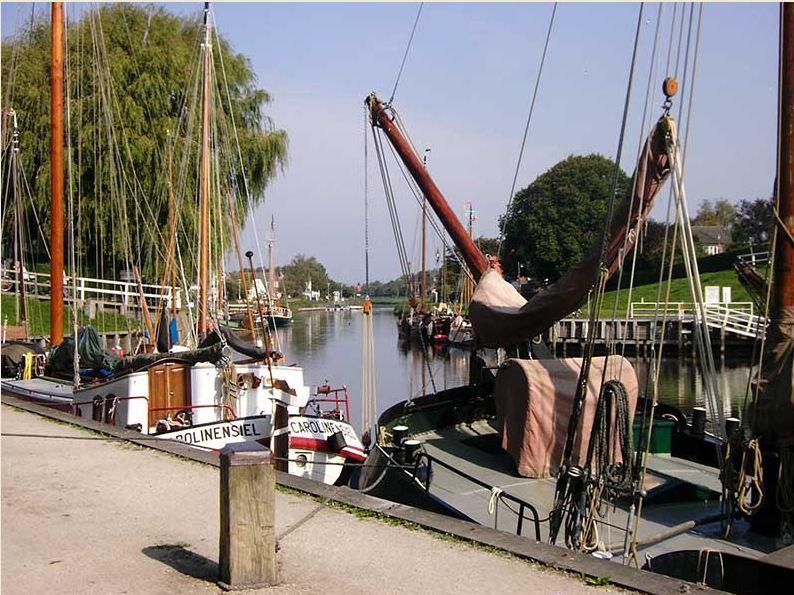 Boats are moored at a harbor with a waterfront path and trees in the background.