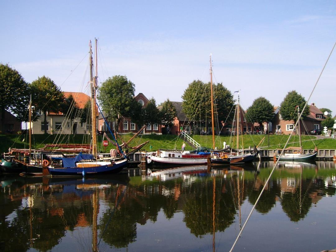 Harbor with several sailboats and reflections in the water. Houses and trees are visible in the background.