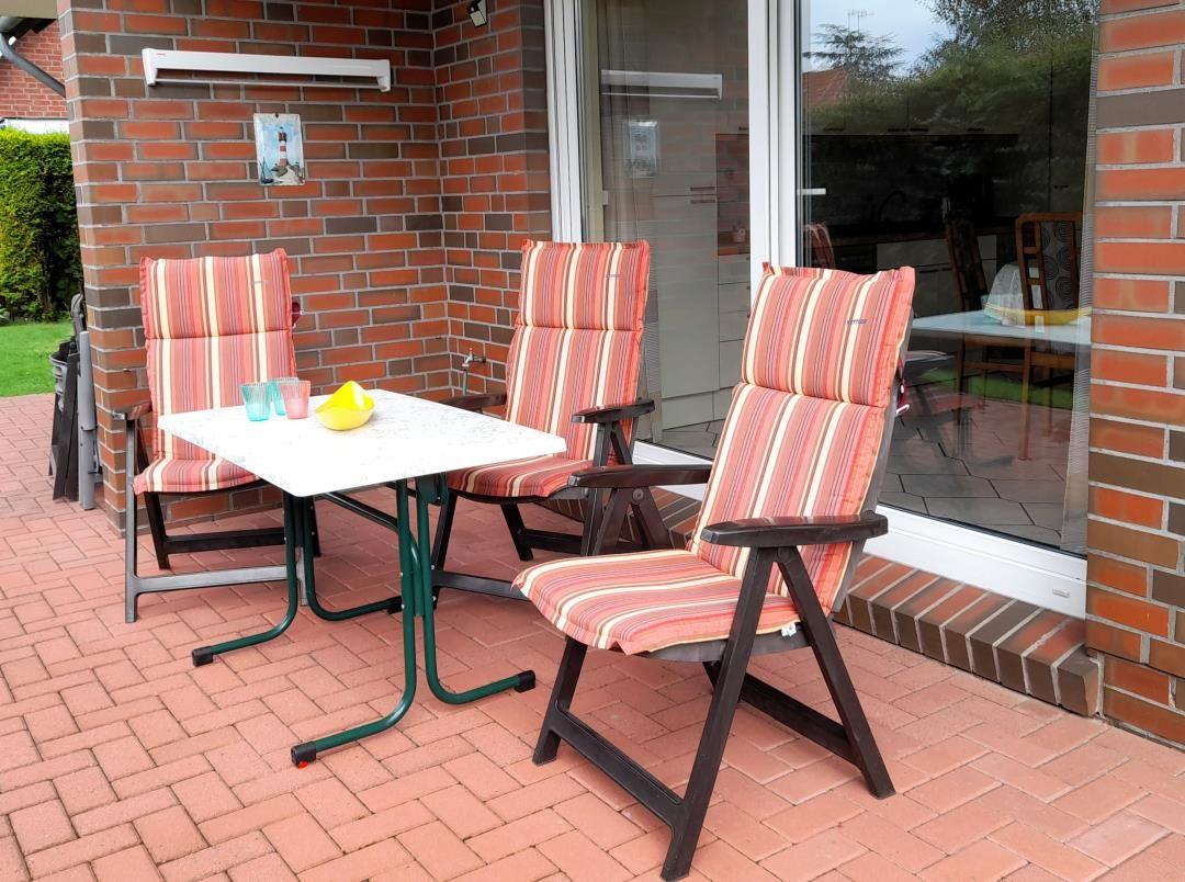 Terrace with table and three chairs with striped cushions. Background: glass door and brick wall.