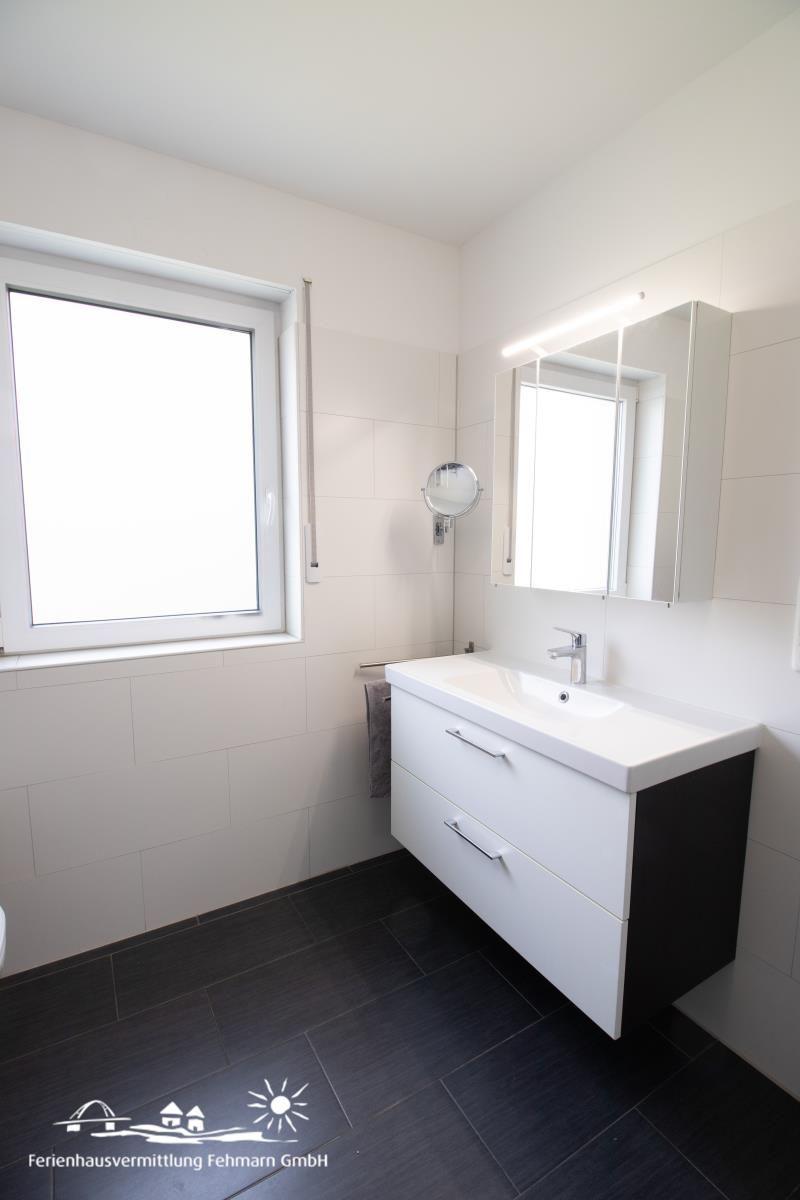 Badezimmer mit Waschbecken, Spiegel und Fenster. Fliesenboden und weiße Wände.