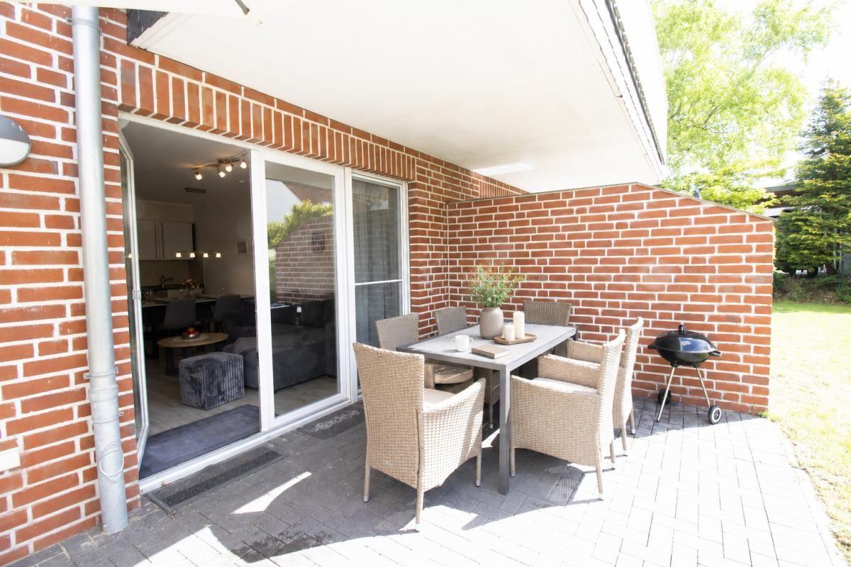 Terrasse mit Tisch, Stühlen und Grill. Durchgang ins Innere. Backsteinmauer und Pflanzen.
