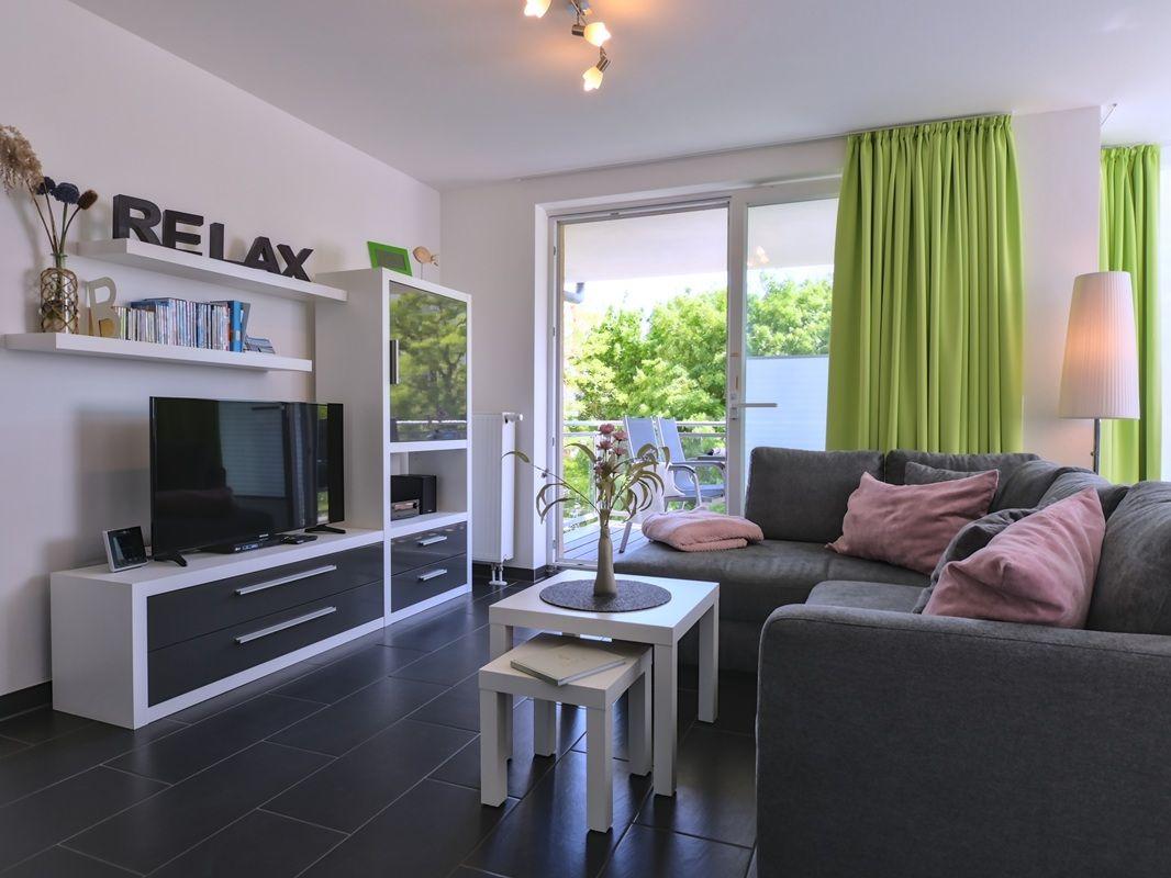 Wohnzimmer mit Sofa, TV und Balkon. Grüne Vorhänge und dunkle Fliesen.
