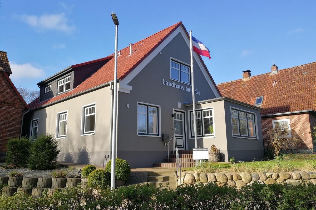 Landhaus am Ufer mit rotem Dach und Flagge. Vorhof mit Steintreppe und Grünflächen.