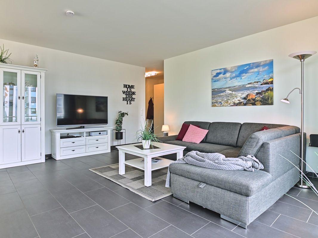 Wohnzimmer mit grauem Sofa, weißem Couchtisch und Fernseher an der Wand.
