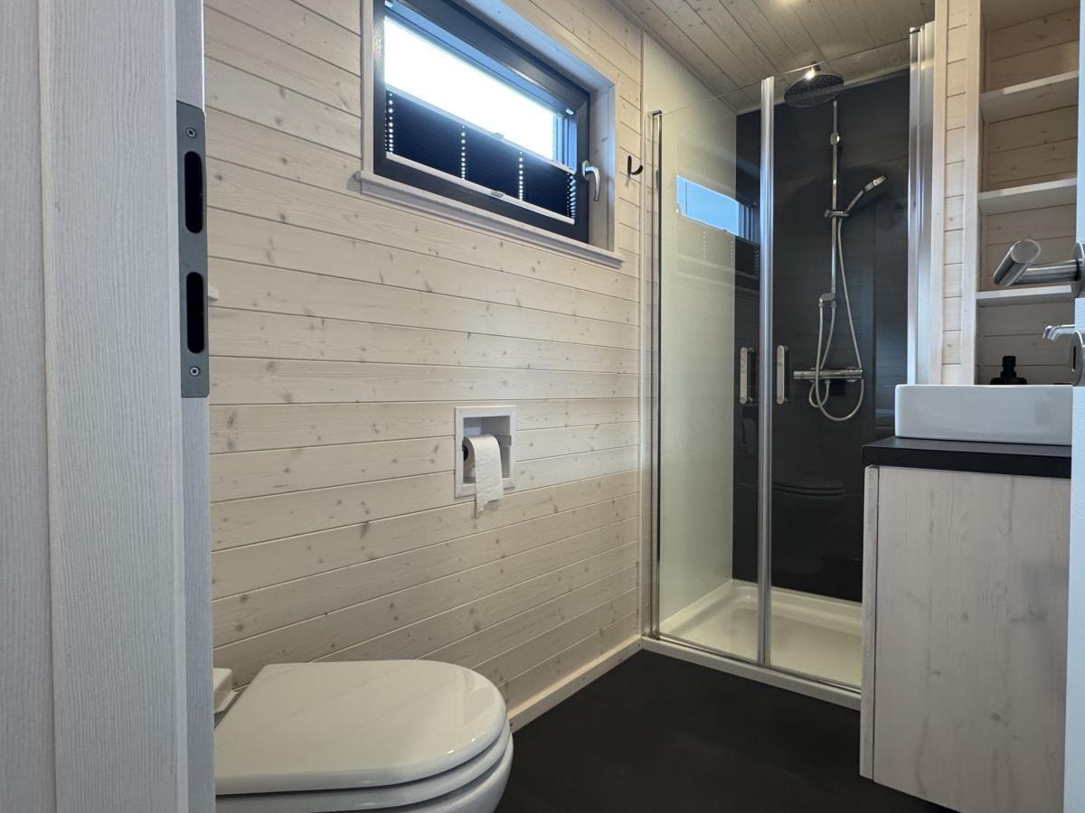 Badezimmer mit Dusche, Waschbecken und Toilette. Holzwand und Fenster.
