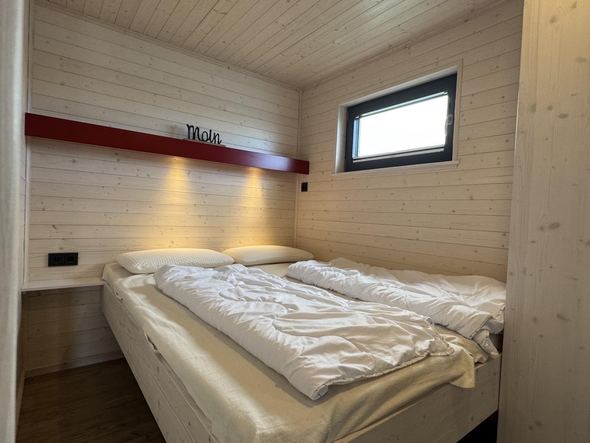 Schlafzimmer mit Bett, Holzwand und Fenster. Über dem Bett steht Moin.