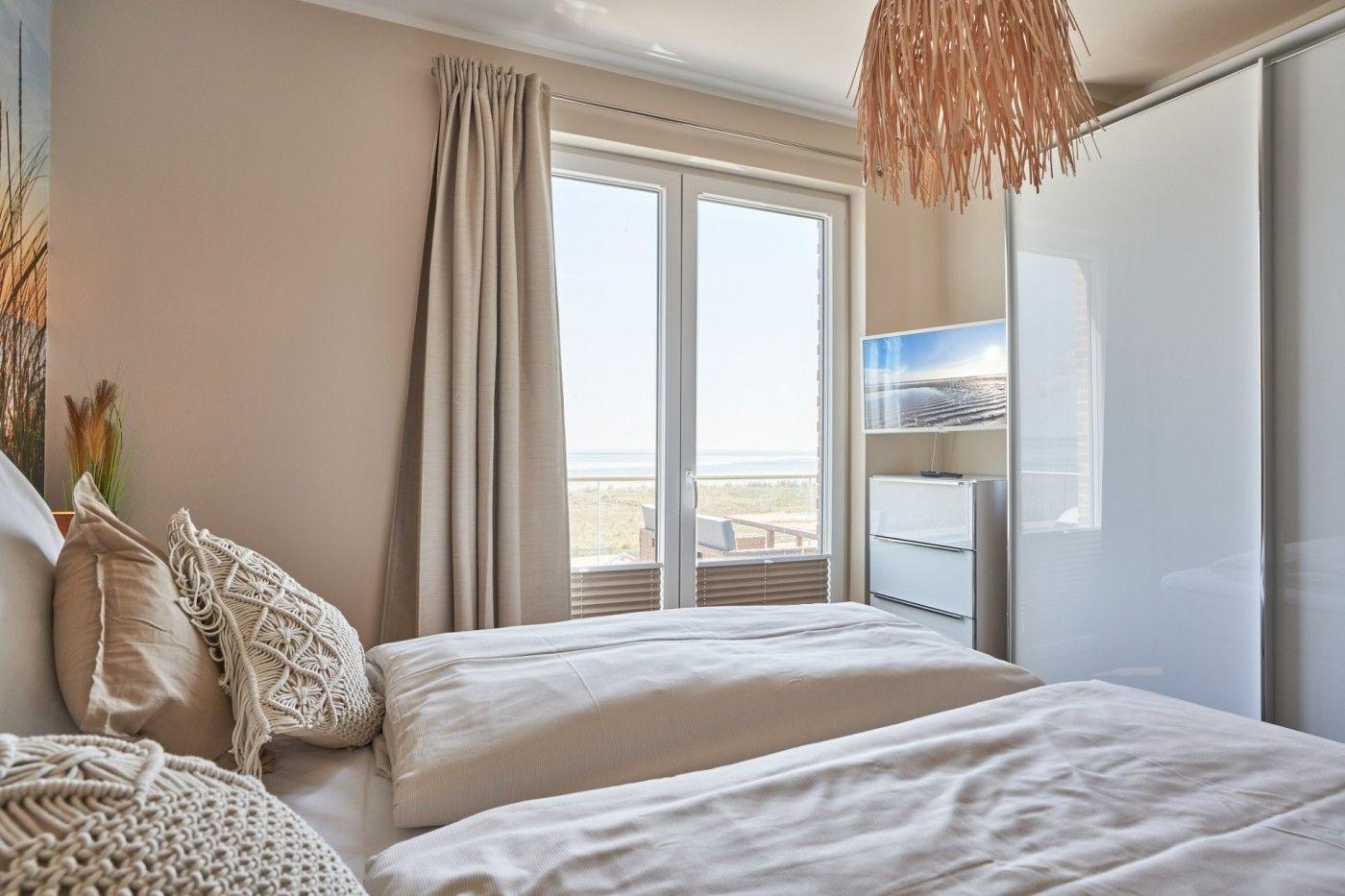 Schlafzimmer mit Blick auf Strand und Meer. Bett, Gardinen und Schrank sind sichtbar.