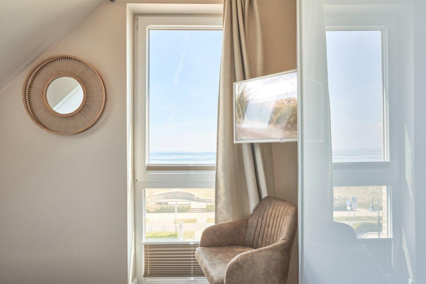 Ein Raum mit Fensterblick aufs Meer, Sessel und runder Wandspiegel.