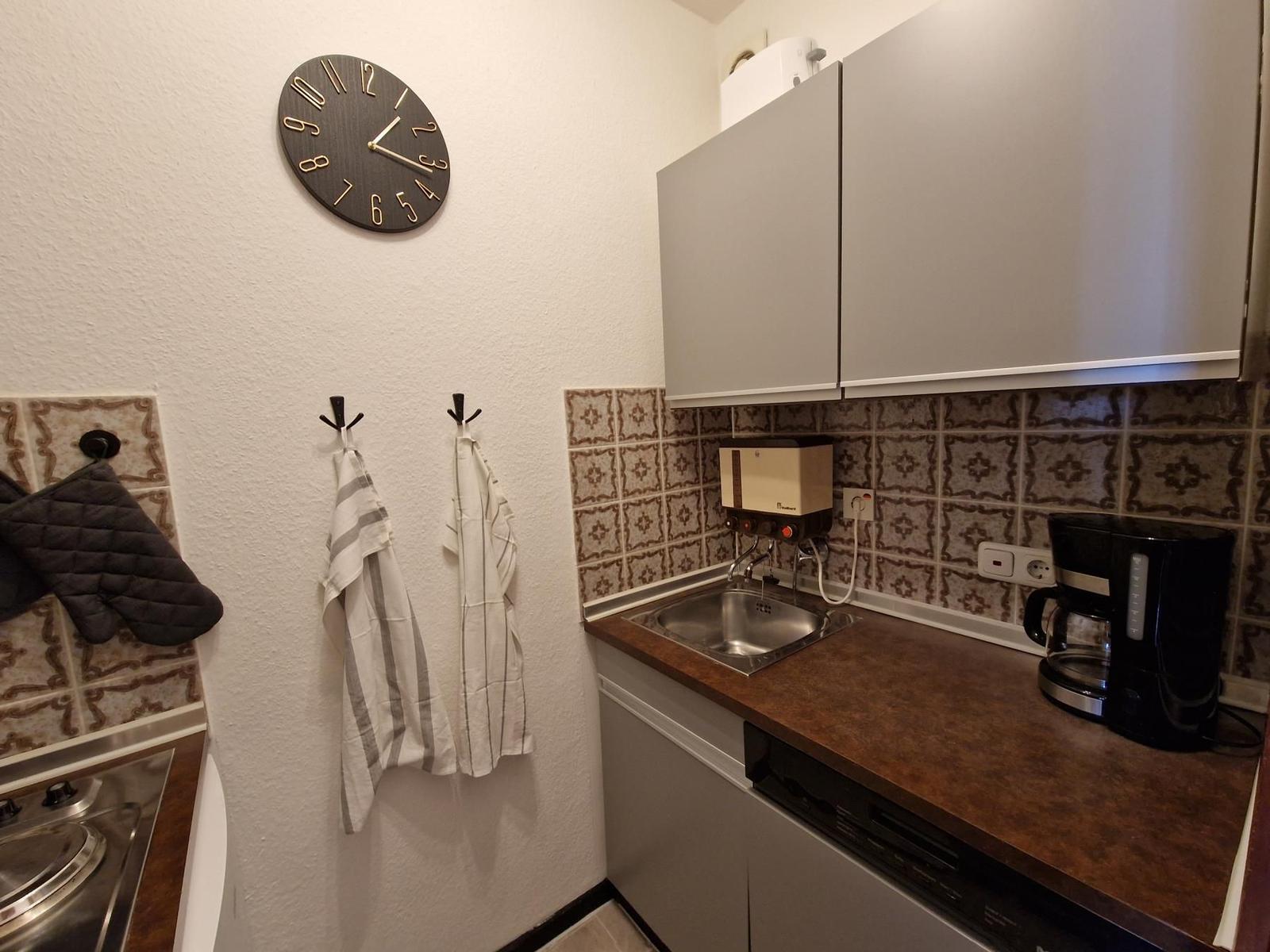 Küche mit Spüle, Kaffeemaschine und Wanduhr. An den Wänden hängen Handtücher und Ofenhandschuhe.