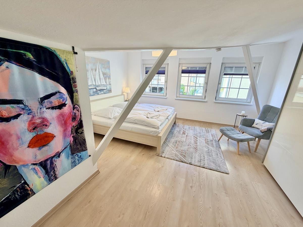 Schlafzimmer mit Bett, Sessel und Wandbild. Holzboden und drei Fenster.