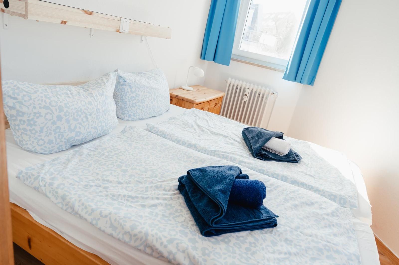 Doppelbett mit blau gemustertem Bettzeug und gefalteten Handtüchern. Holz-Kopfteil und Nachttisch. Blaue Vorhänge.