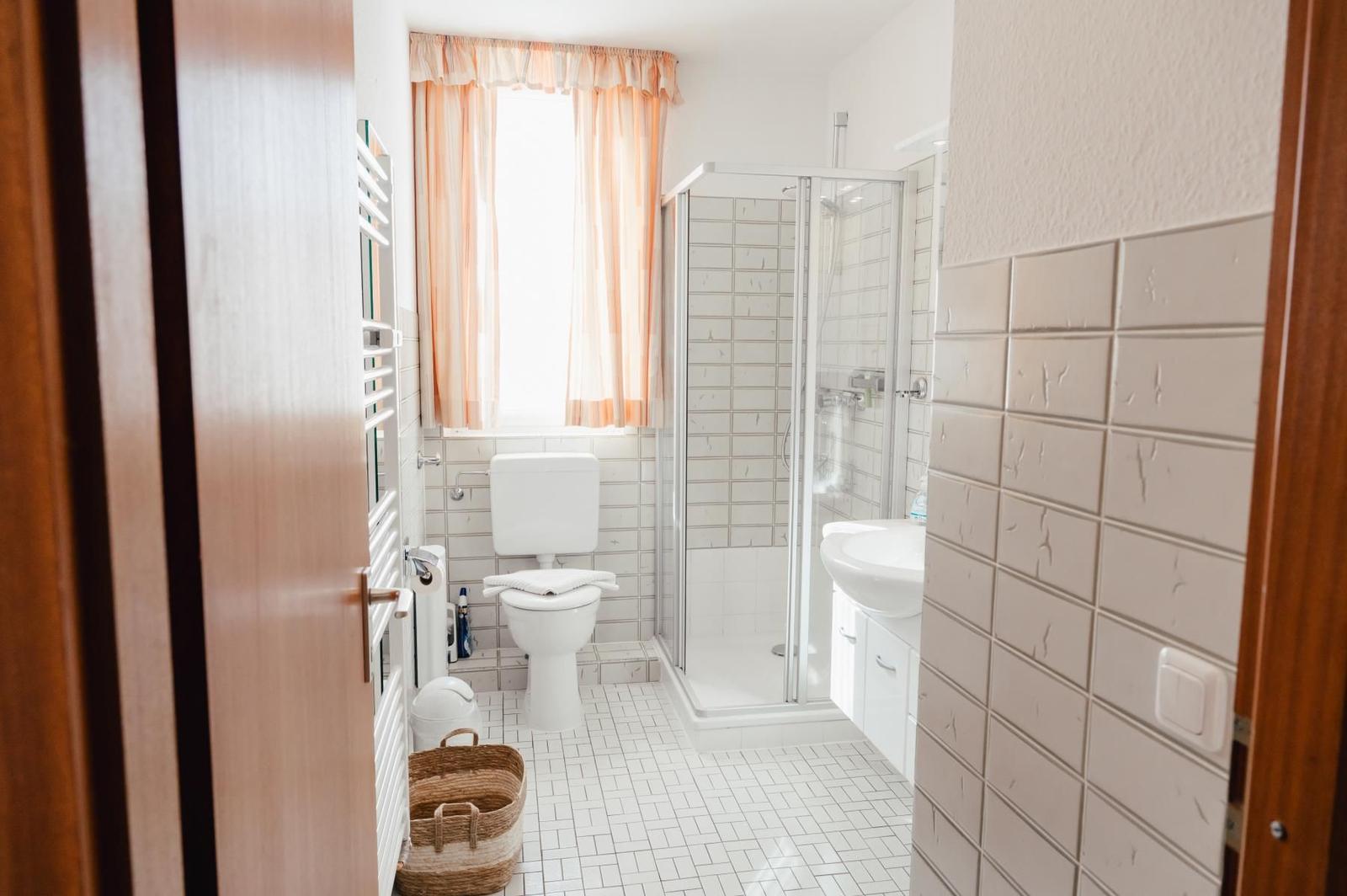 Badezimmer mit Dusche, Toilette und Waschbecken. Fenster mit Vorhängen. Fliesenboden und Wandfliesen.