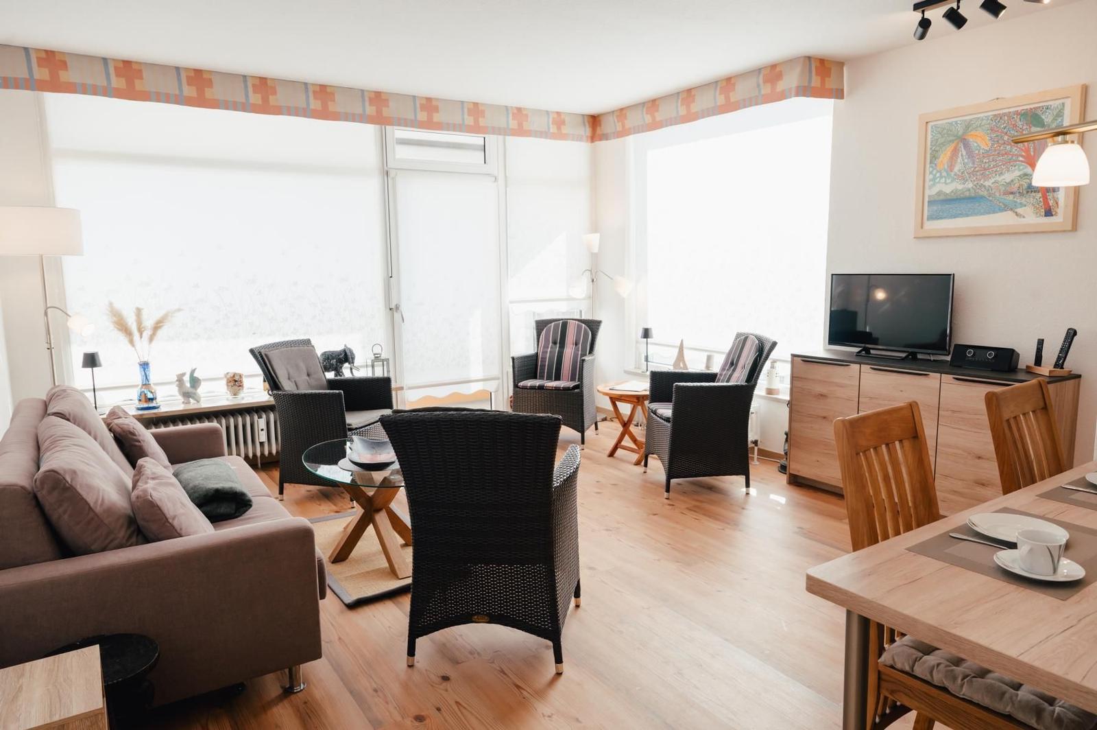 Wohn- und Essbereich mit Sofa, Stühlen und TV. Holzboden und große Fenster.