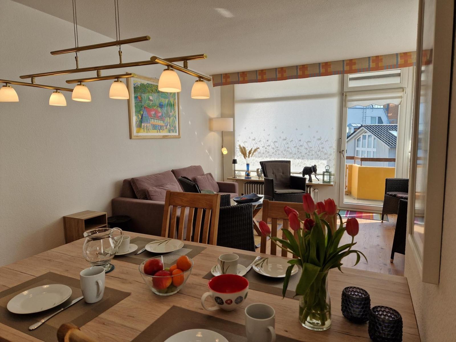 Essbereich mit Tisch, Stühlen und Blumen. Im Hintergrund Wohnzimmer mit Sofa und Balkon.