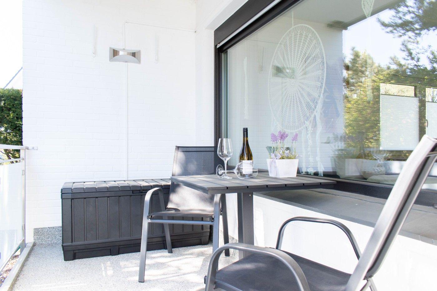 Terrasse mit Tisch, Stühlen und Wein. Hinter Glas ist ein Traumfänger sichtbar.