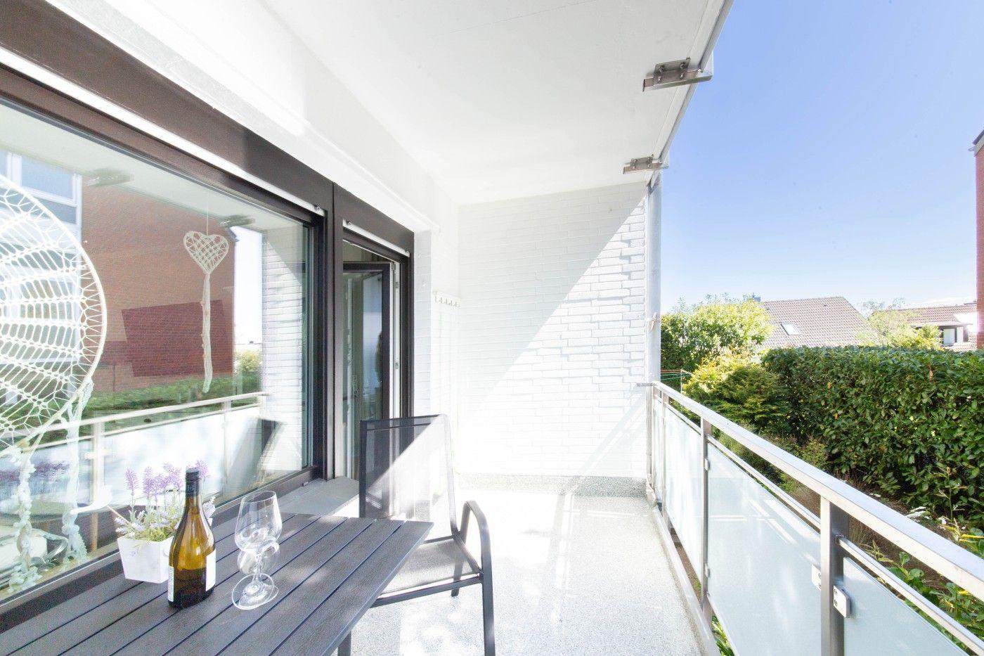 Terrasse mit Tisch, Stuhl und Weinflasche. Glasgeländer und Blick auf grüne Hecke.