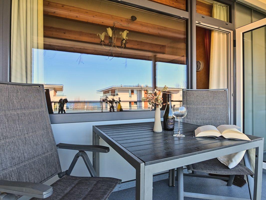 Terrasse mit Tisch, zwei Stühlen, Wein und Buch. Blick auf Wohnhaus und See.