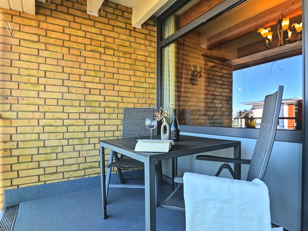 Terrasse mit Tisch, zwei Stühlen, Flasche und Glas. Gelbe Ziegelwand und Glasfront.