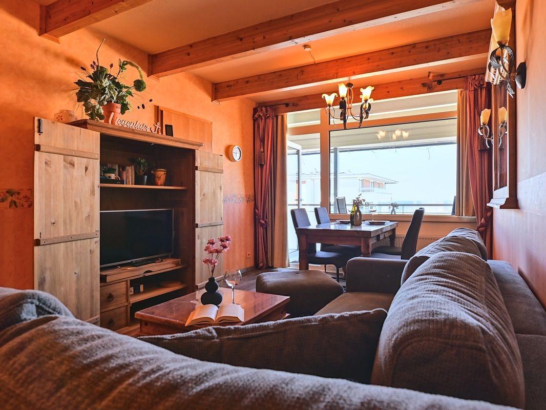 Wohn- und Essbereich mit Sofa, Fernseher und großem Fenster. Holzdecke und warme Farben.