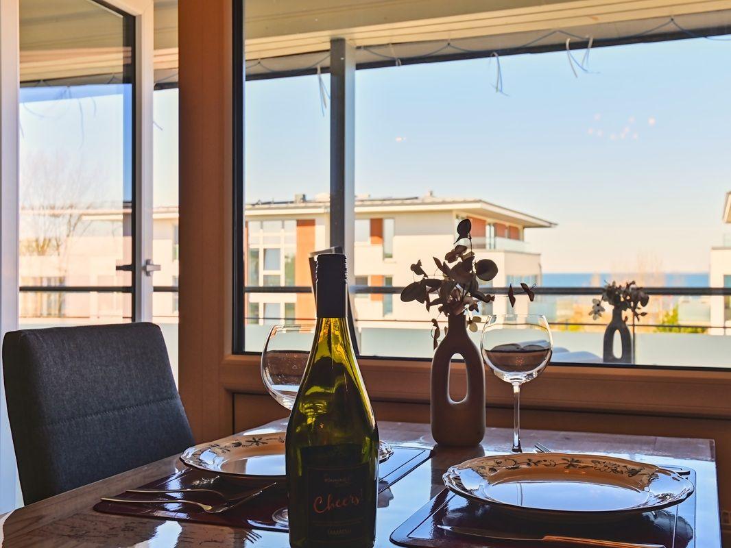 Tisch mit Weinflasche und Gläsern vor großem Fenster mit Blick auf Gebäude und Meer.