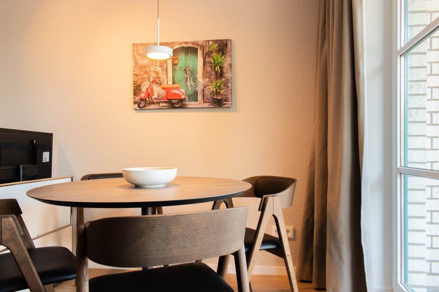 Runder Tisch mit Stühlen, Wandbild und Fenster mit Vorhang.