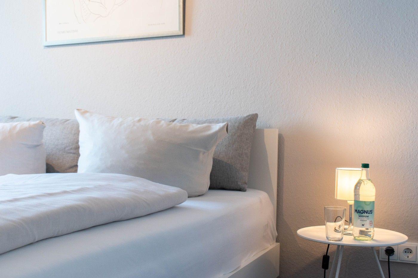 Schlafzimmer mit Bett, Kissen und Nachttisch mit Lampe und Wasserflasche.