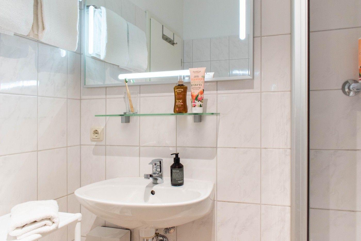 Badezimmer mit Waschbecken, Spiegel und Dusche. Auf der Ablage stehen Seife und Sonnencreme.