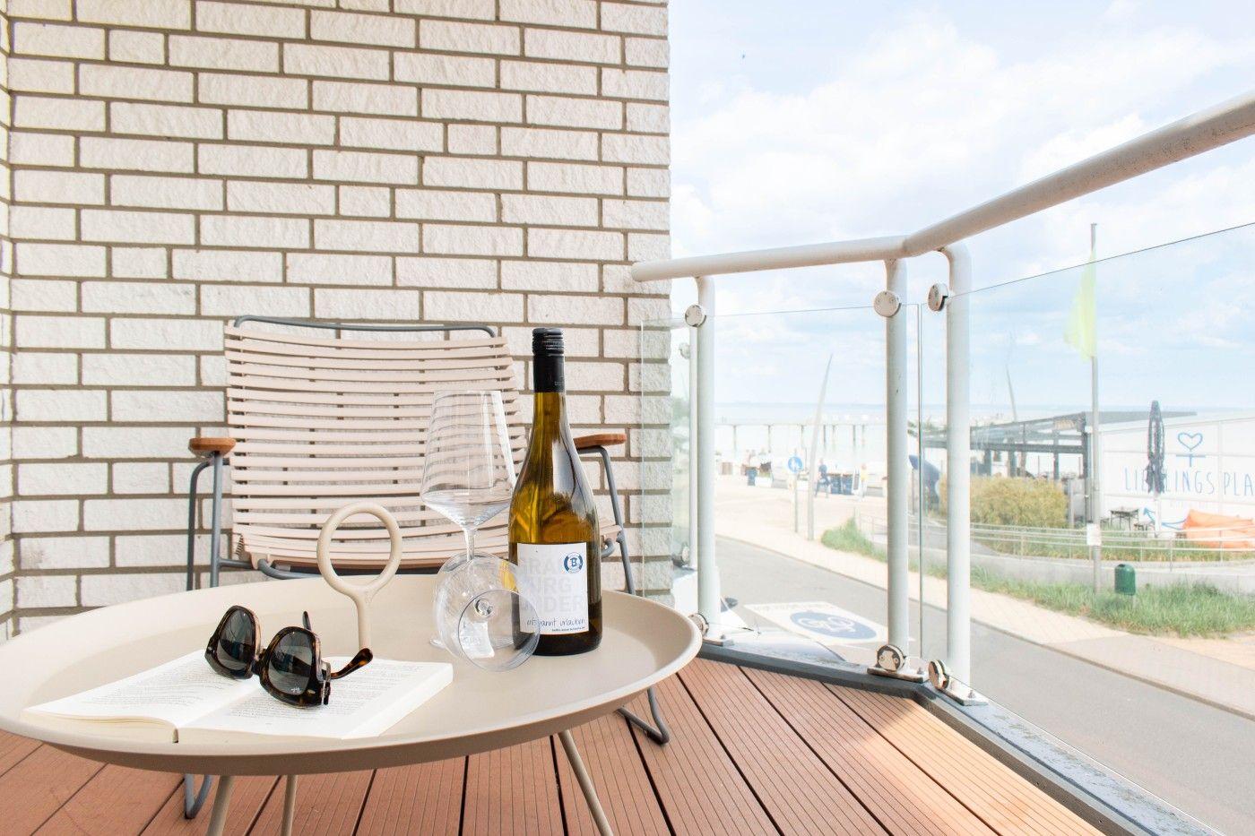 Terrasse mit Tisch, Stuhl, Weinflasche und Sonnenbrille. Blick auf Hafen und Strand.