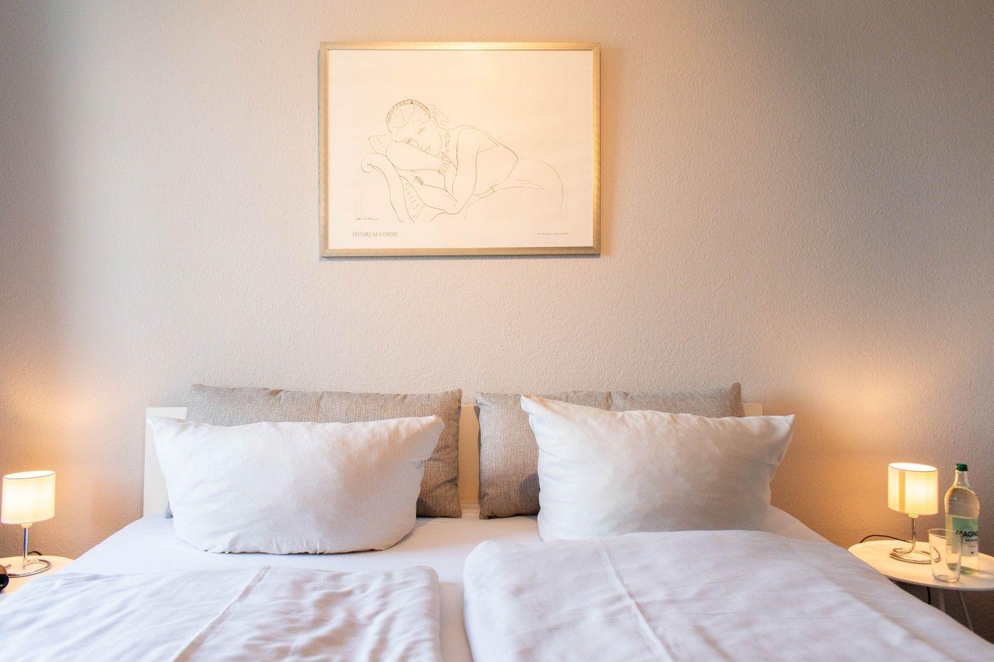 Ein Bett mit weißen Kissen und einer Decke. Über dem Bett hängt ein gerahmtes Kunstwerk.