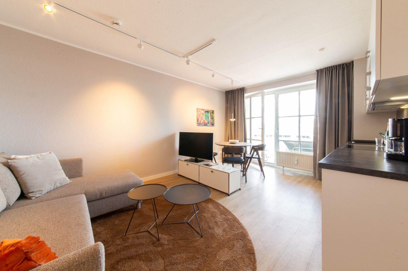 Wohnung mit Sofa, TV, Esstisch und Balkon. Küche mit Arbeitsplatte und Kaffeemaschine.