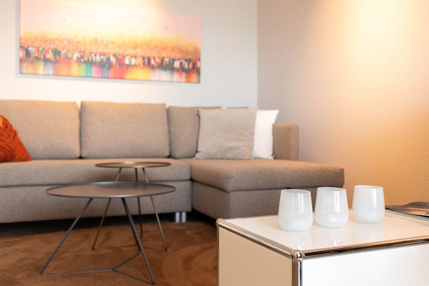 Ein Wohnzimmer mit einem grauen Sofa, zwei runden Tischen und einer weißen Kommode mit drei Gläsern.