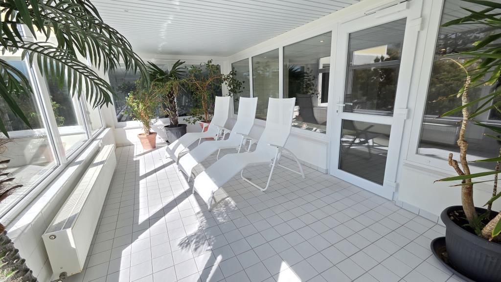 Terrasse mit weißen Liegestühlen und Pflanzen. Durchgehende Glasfront und weißer Fliesenboden.