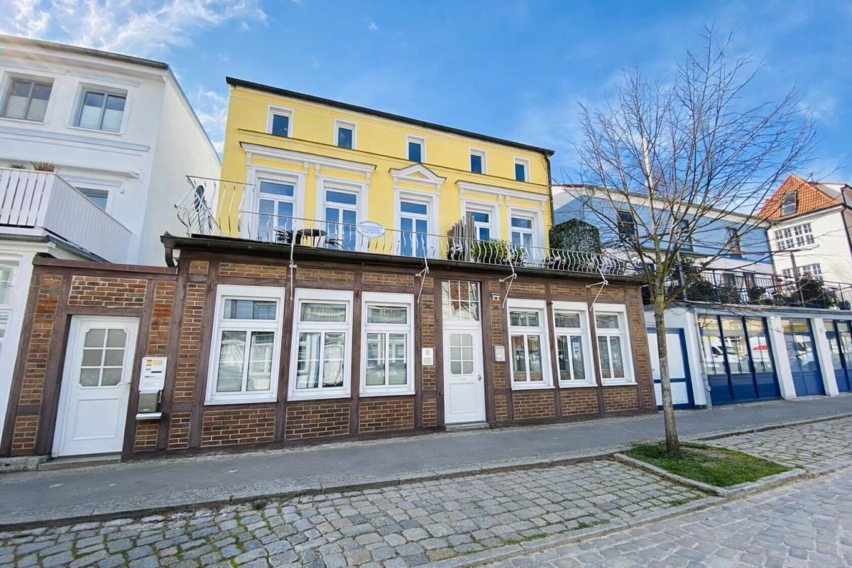 Gelbes Haus mit Balkon und rotem Backsteinbau an Straße mit Kopfsteinpflaster