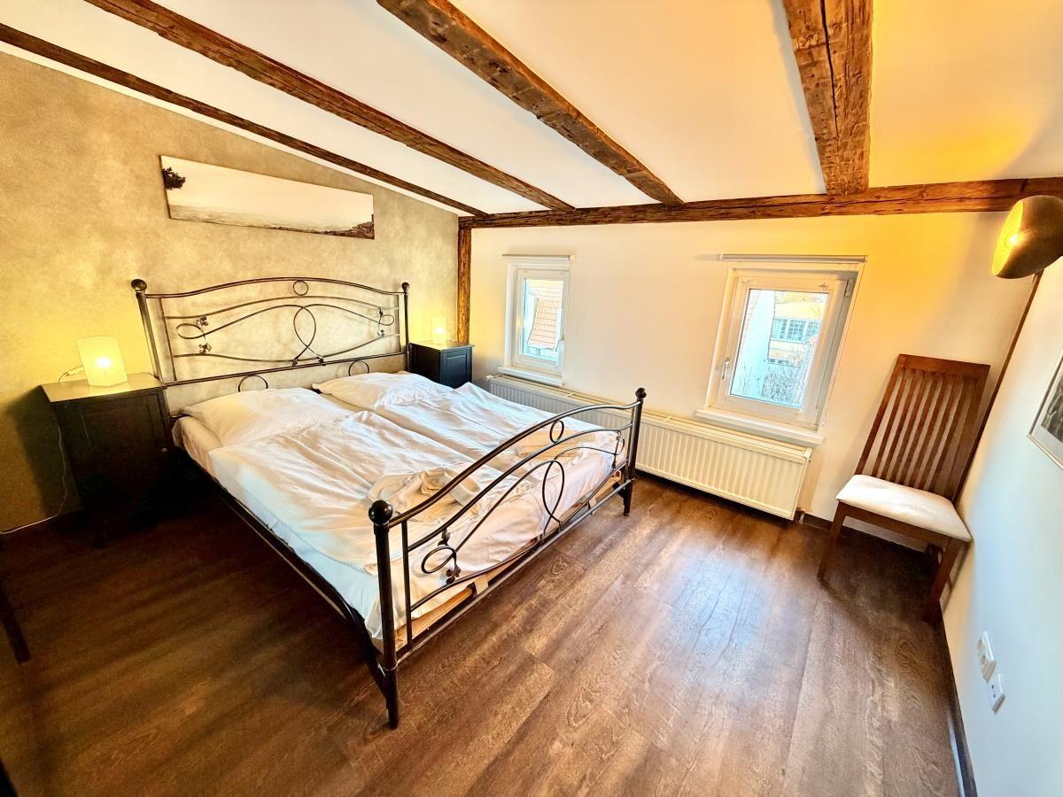 Schlafzimmer mit Doppelbett, Holzboden und Holzbalkendecke. Zwei Fenster und Stuhl.