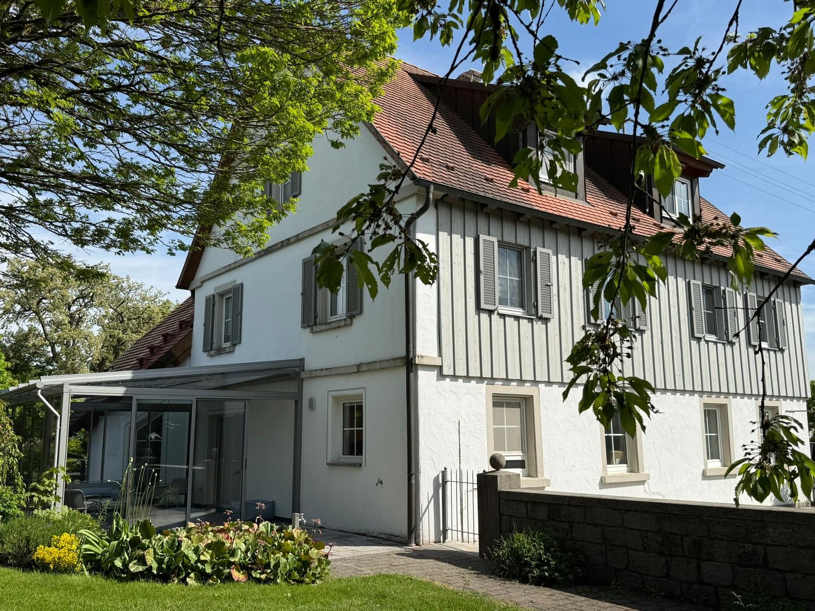 Ein zweistöckiges Haus mit weißer Fassade, Holzverkleidung und rotem Dach. Garten und Terrasse sind sichtbar.