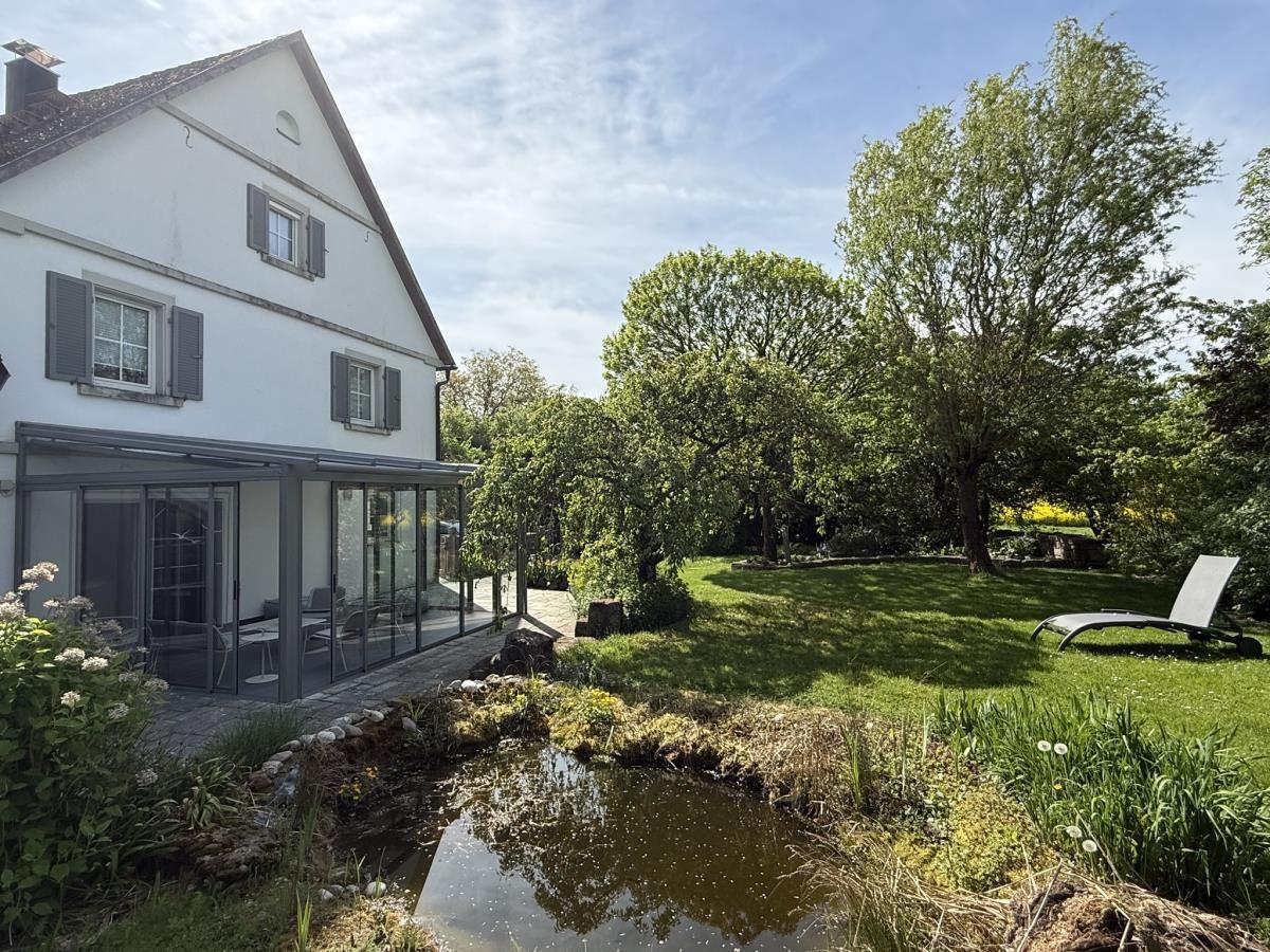 Haus mit Garten, Teich und Liegestuhl unter Bäumen.