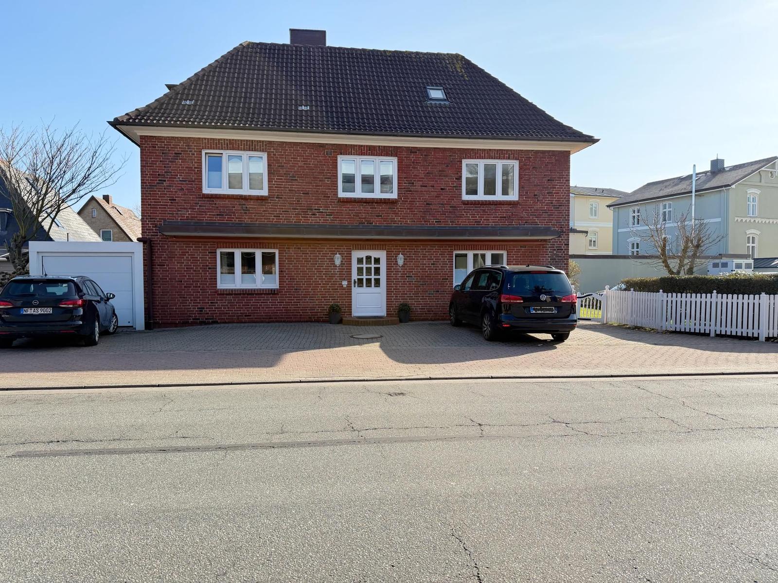 Zweistöckiges Haus mit rotem Backsteinfassade, weißer Tür und Fenstern. Zwei Autos parken auf dem Hof.