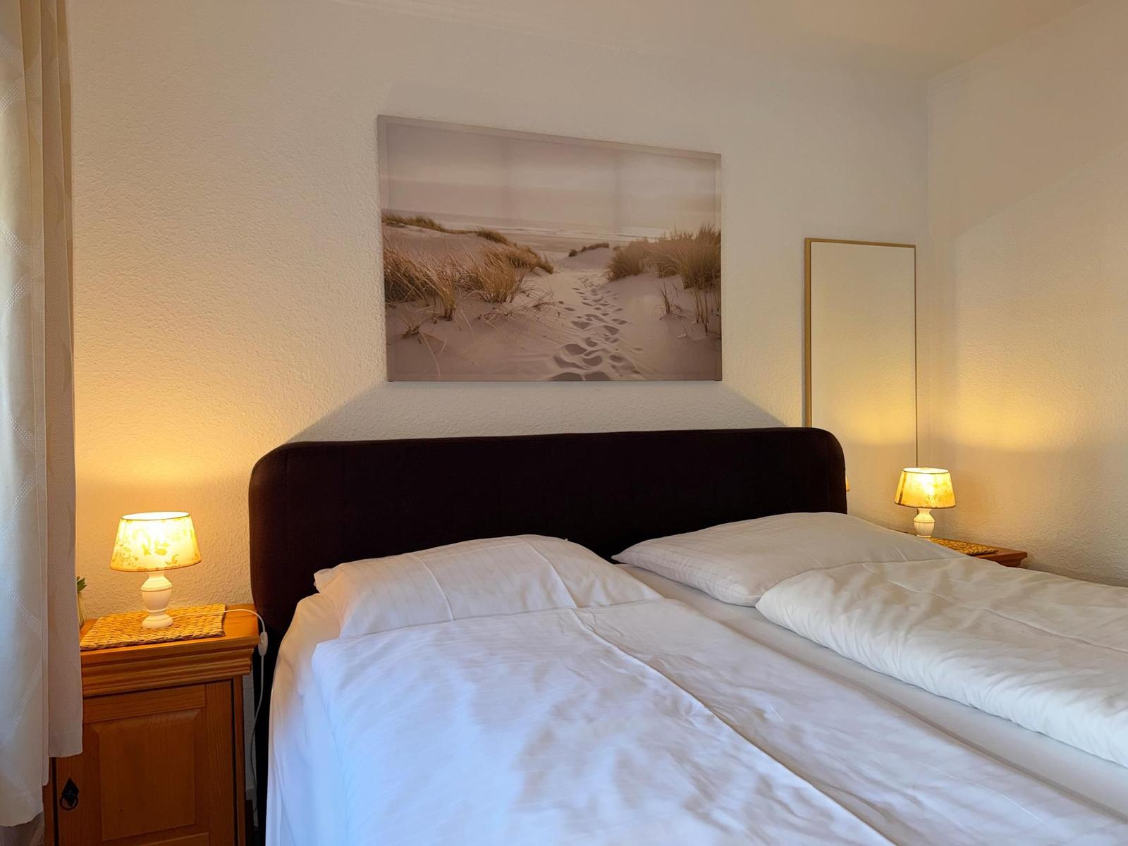 Schlafzimmer mit Bett, Nachttischen und Wandbild. Beleuchtung durch Lampen.