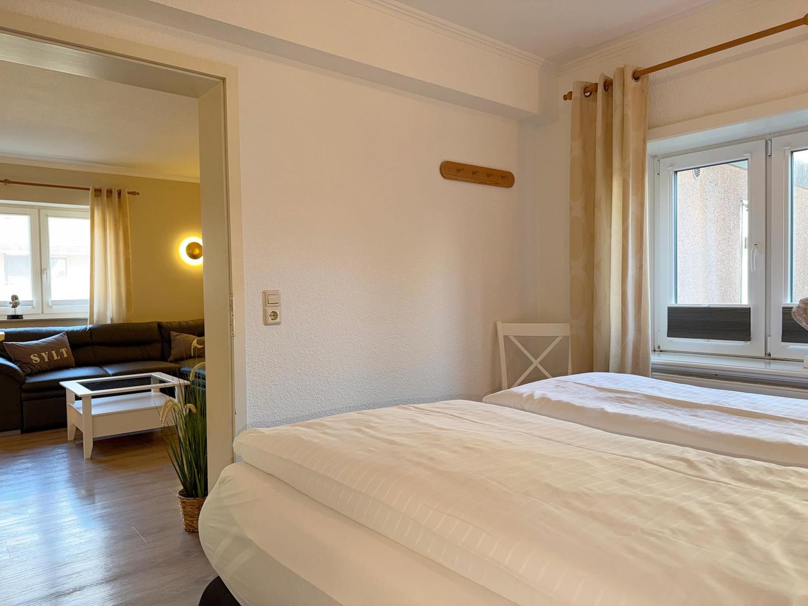 Zwei Betten mit weißen Bettwäsche. Durchgang in Wohnzimmer mit Sofa und Fenster.