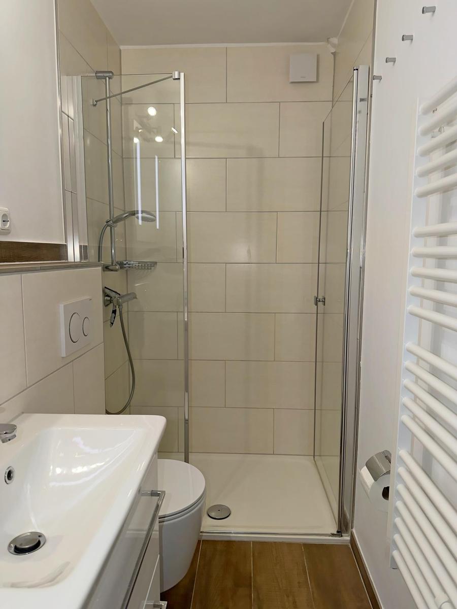 Bad mit Dusche, Waschbecken und Toilette. Holzdielenboden und weiße Fliesen.