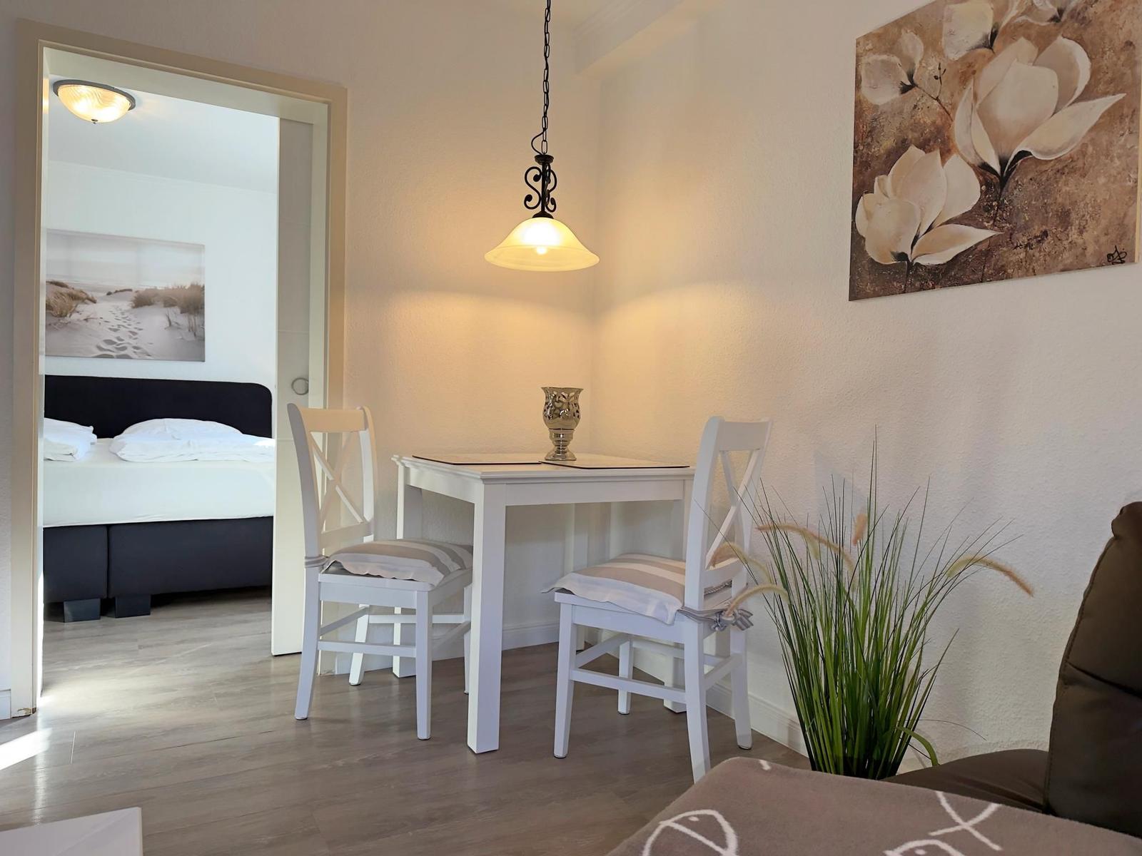 Essbereich mit weißem Tisch und Stühlen, Wandbild und Pflanze. Schlafzimmer mit Bett im Hintergrund.