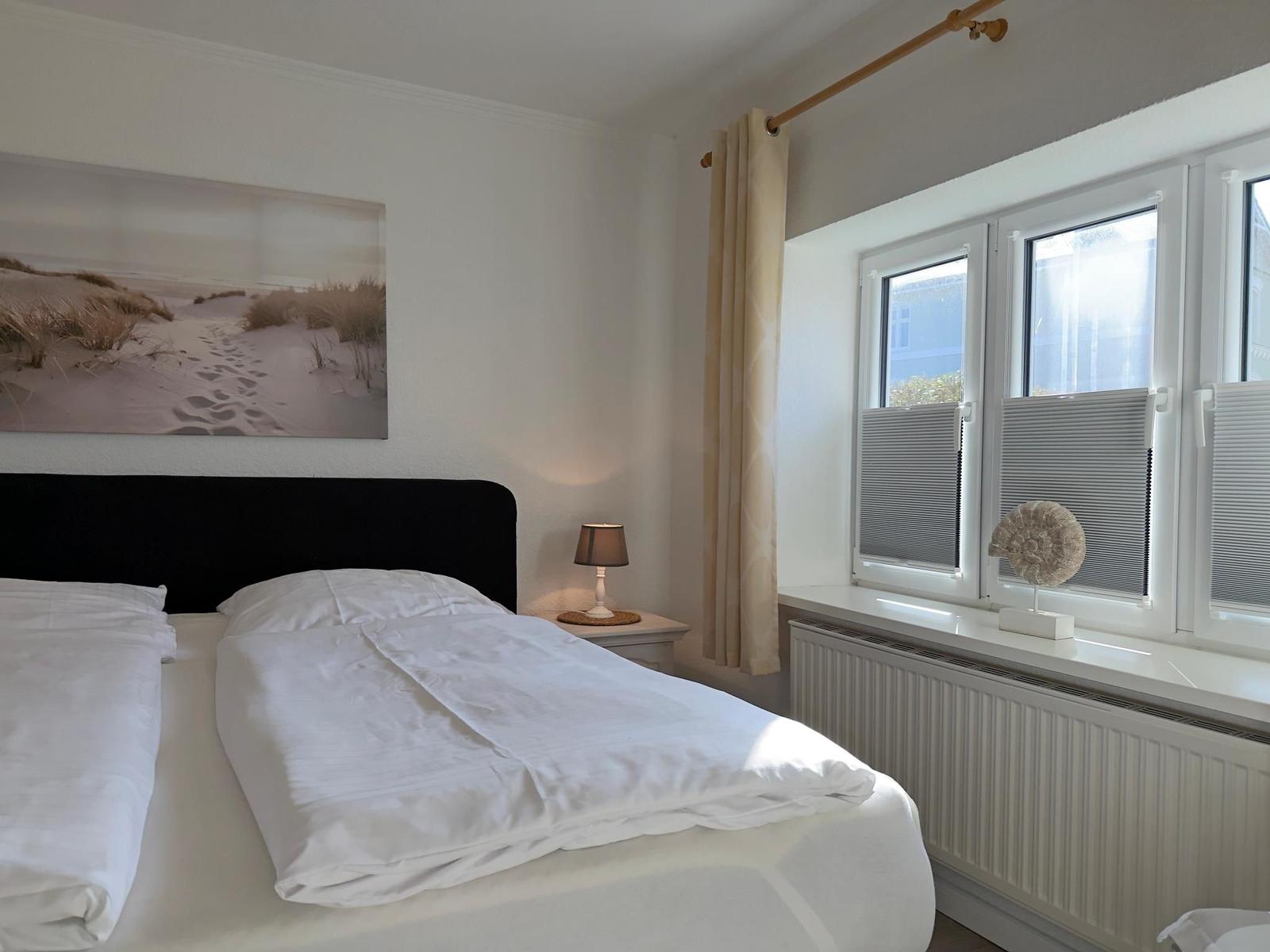 Schlafzimmer mit Bett, Nachttisch und Fenster mit Jalousien.