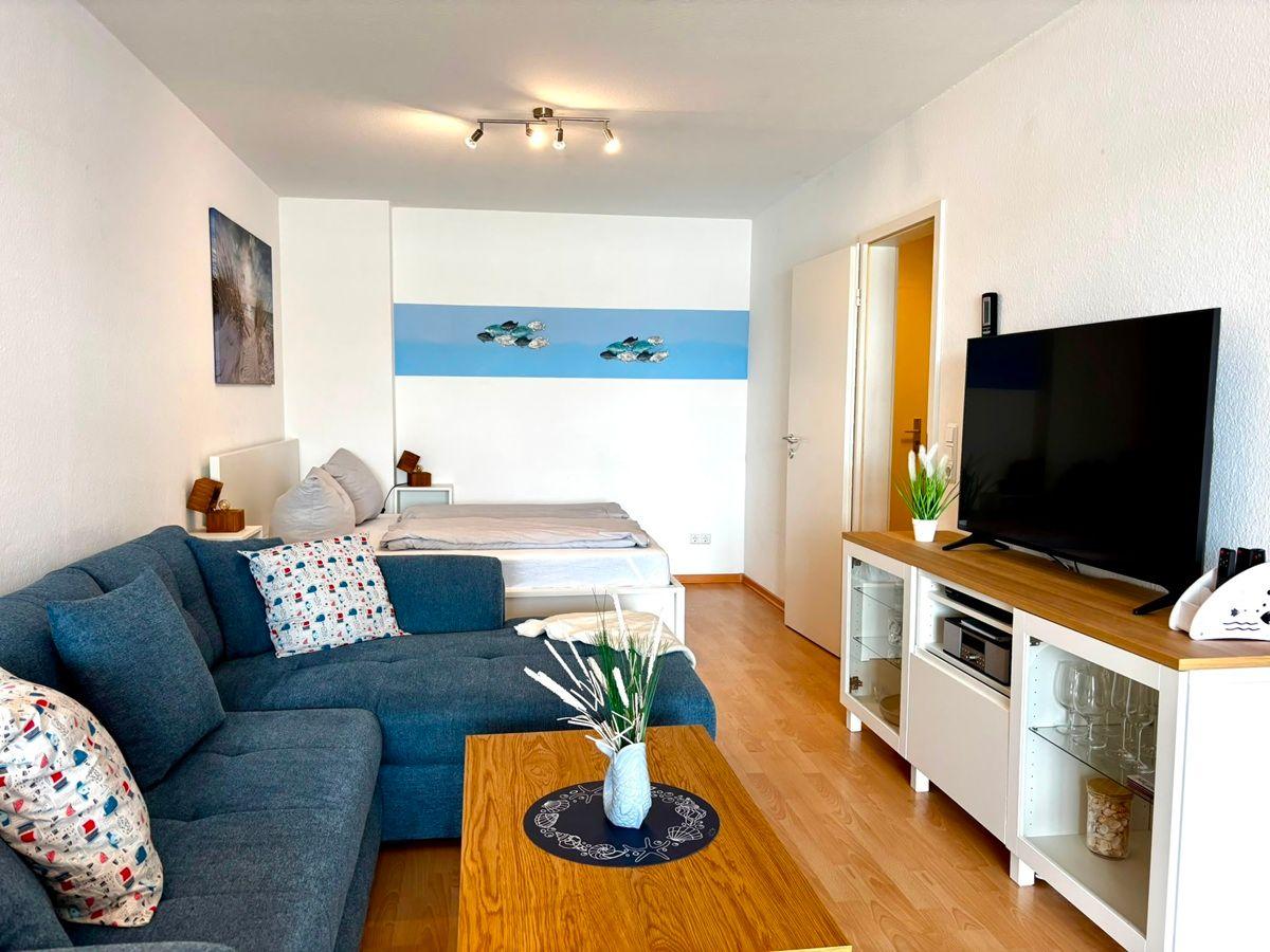Wohnzimmer mit Sofa, Couchtisch und Fernseher. Hintergrund: Schlafbereich mit Bett.
