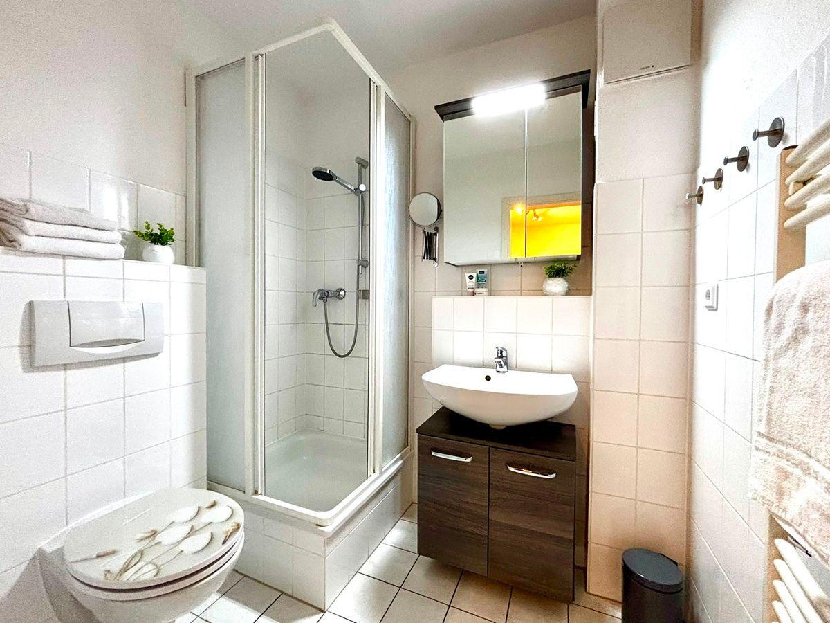 Badezimmer mit Dusche, Waschbecken und Toilette. Weiße Fliesen und Holzschränke.