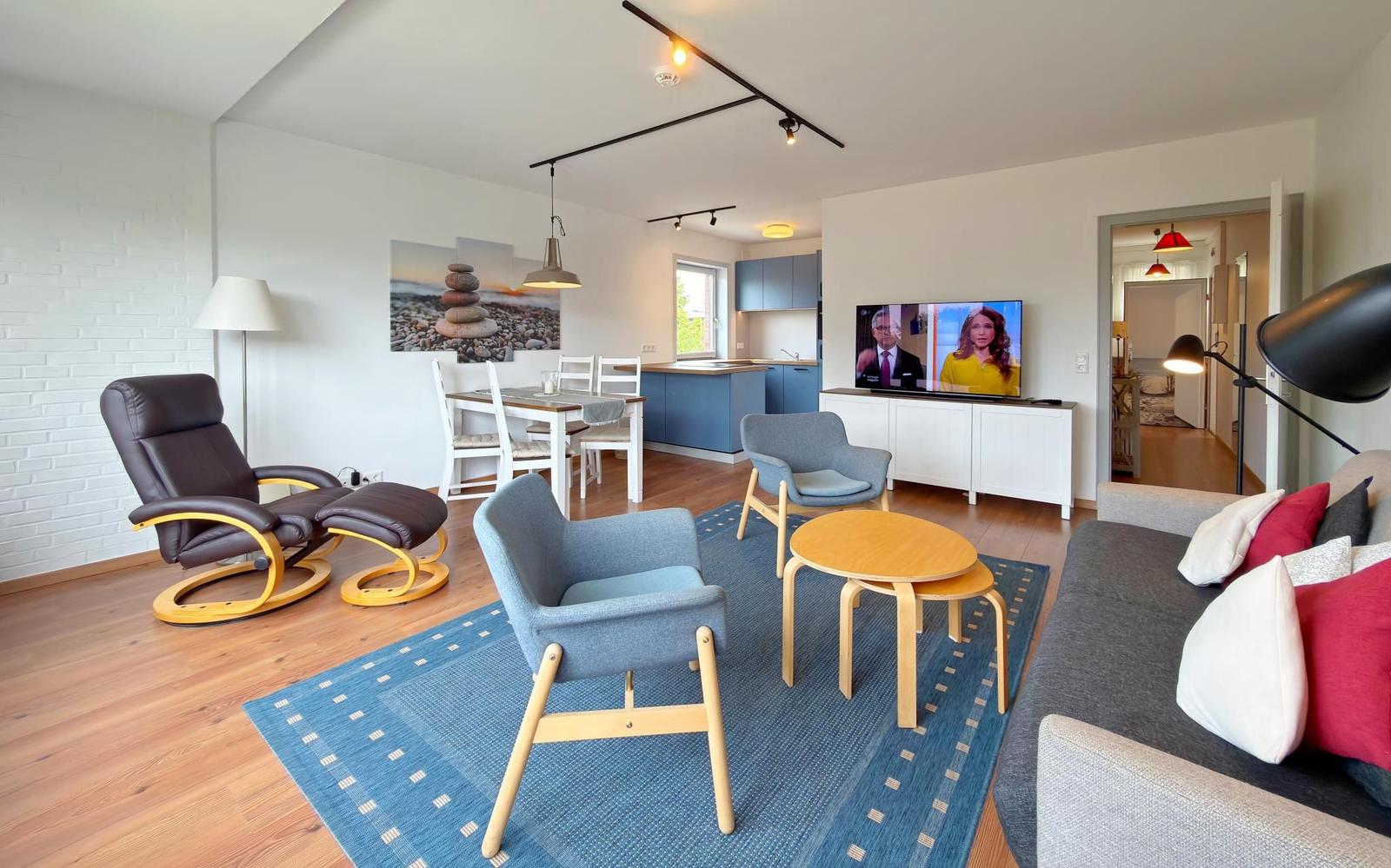 Wohnbereich mit Sofa, Sesseln und Fernseher. Küche und Essbereich im Hintergrund.