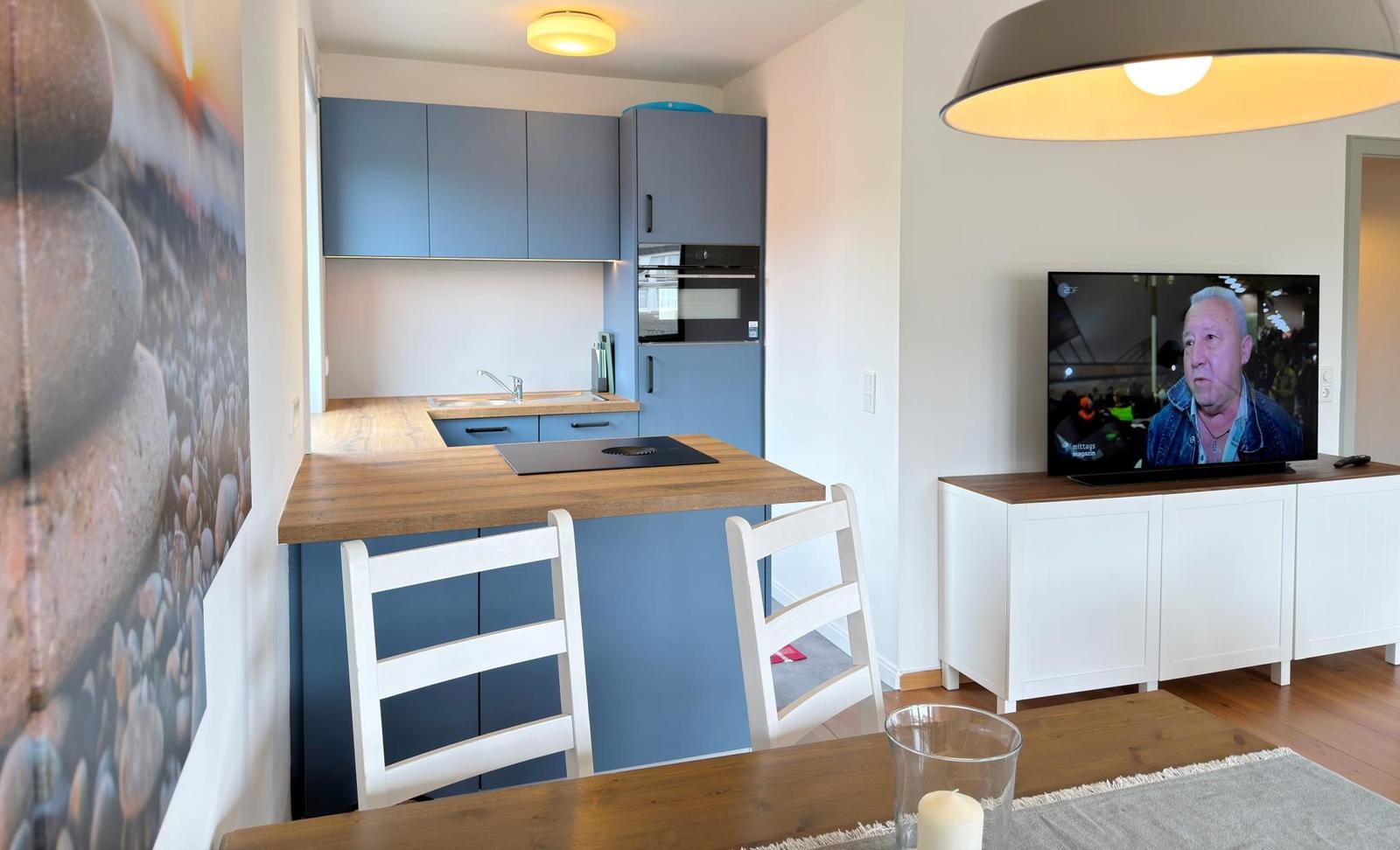 Küche mit Arbeitsplatte und zwei Stühlen. Fernseher auf weißen Schrank. Wandbild mit Steinen.